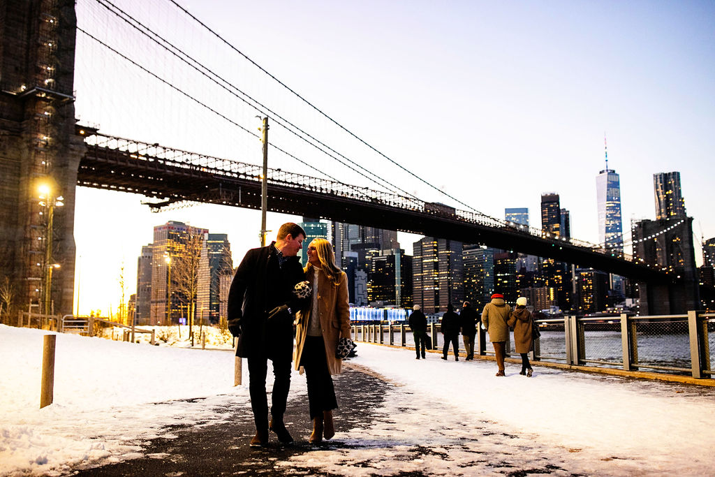 Couple walking under the bridge with the Manhattan skyline in the background during their Brooklyn engagement session during the winter time.
