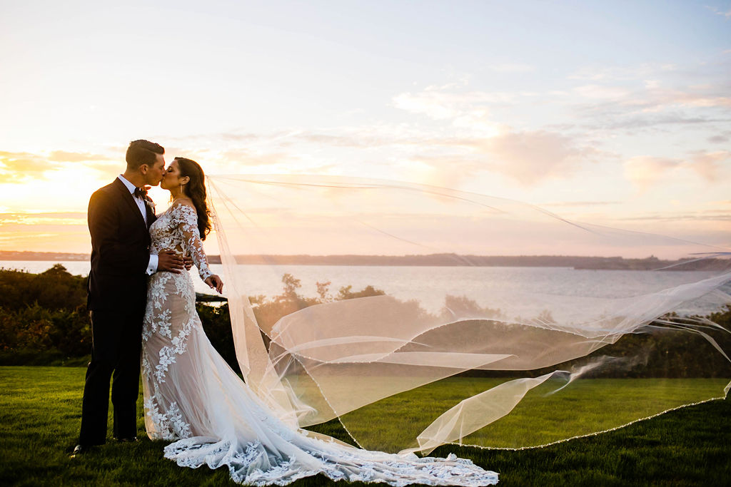 Bride and Groom kiss at sunset with her veil blowing in the wind on the lawn of their Oceancliff wedding in Newport