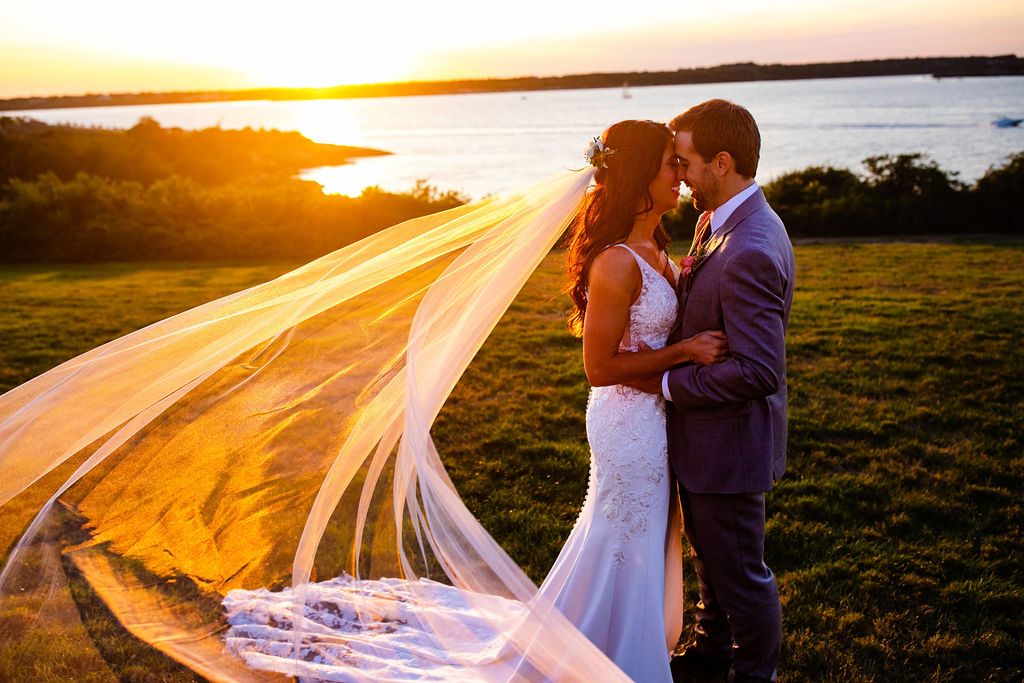 Bride and Groom kiss at sunset with her veil blowing in the wind on the lawn of their Oceancliff wedding in Newport