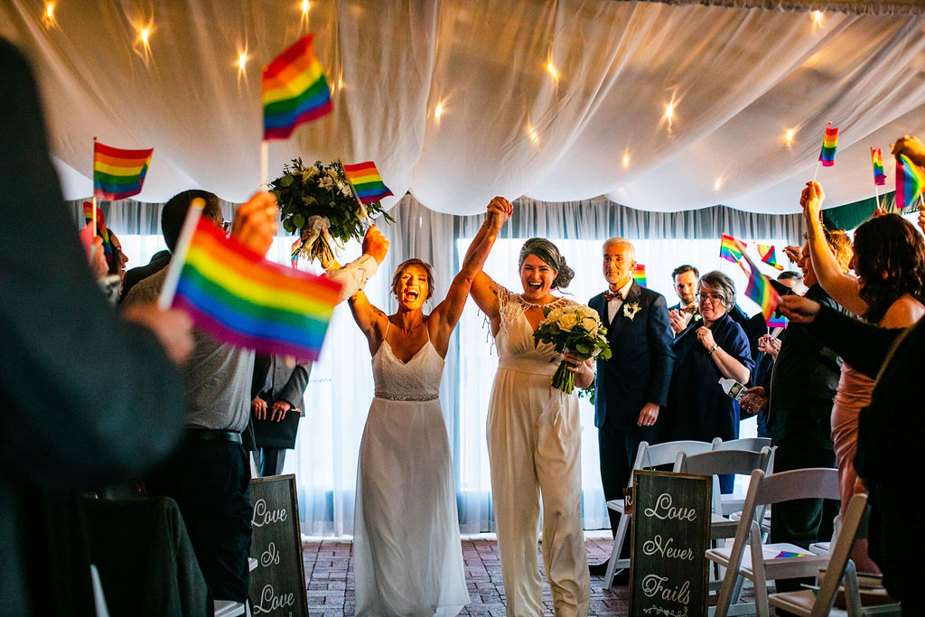 Guests wave rainbow pride flags as two brides walk down the aisle after their wedding ceremony