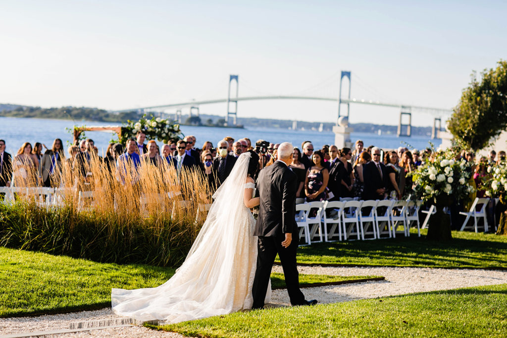 Bride walks with her father to her wedding ceremony on the water with the Newport Bridge in the background