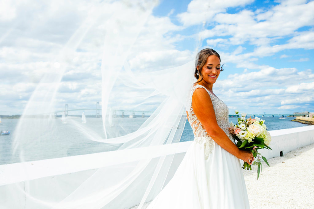 Bride with veil blowing in the wind on a beautiful blue sky day with the newport bridge behind them at Belle Mer