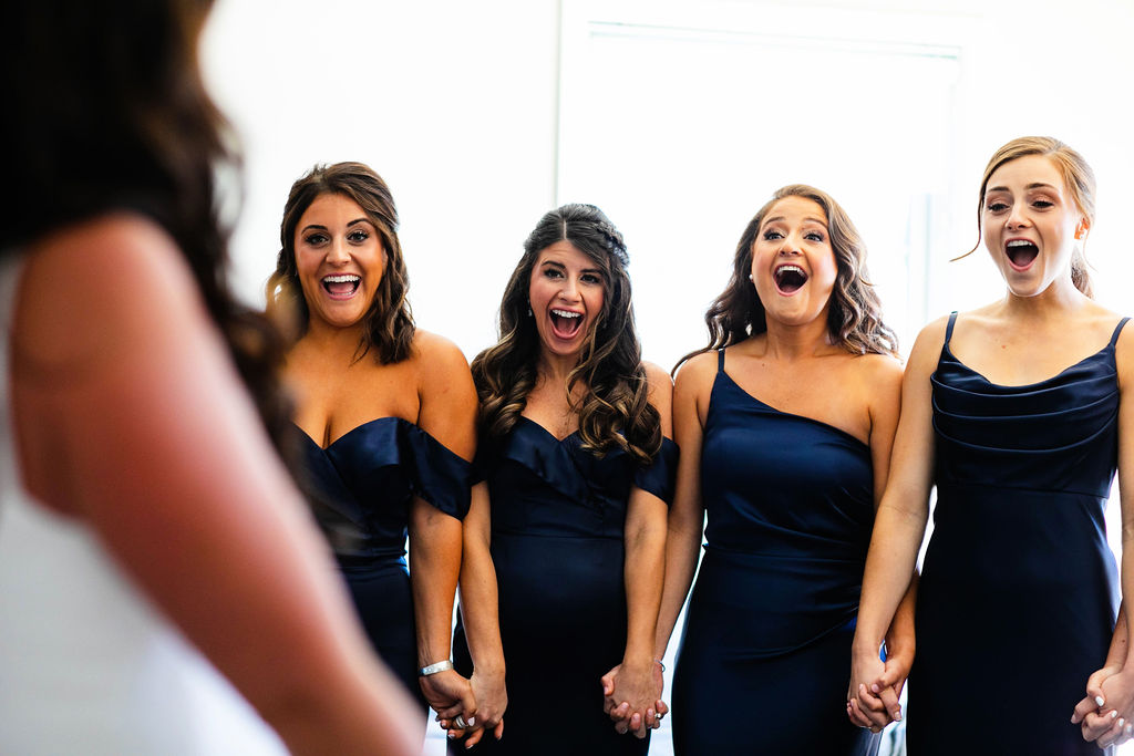 Bridesmaids in navy blue dresses react to seeing the bride for the first time