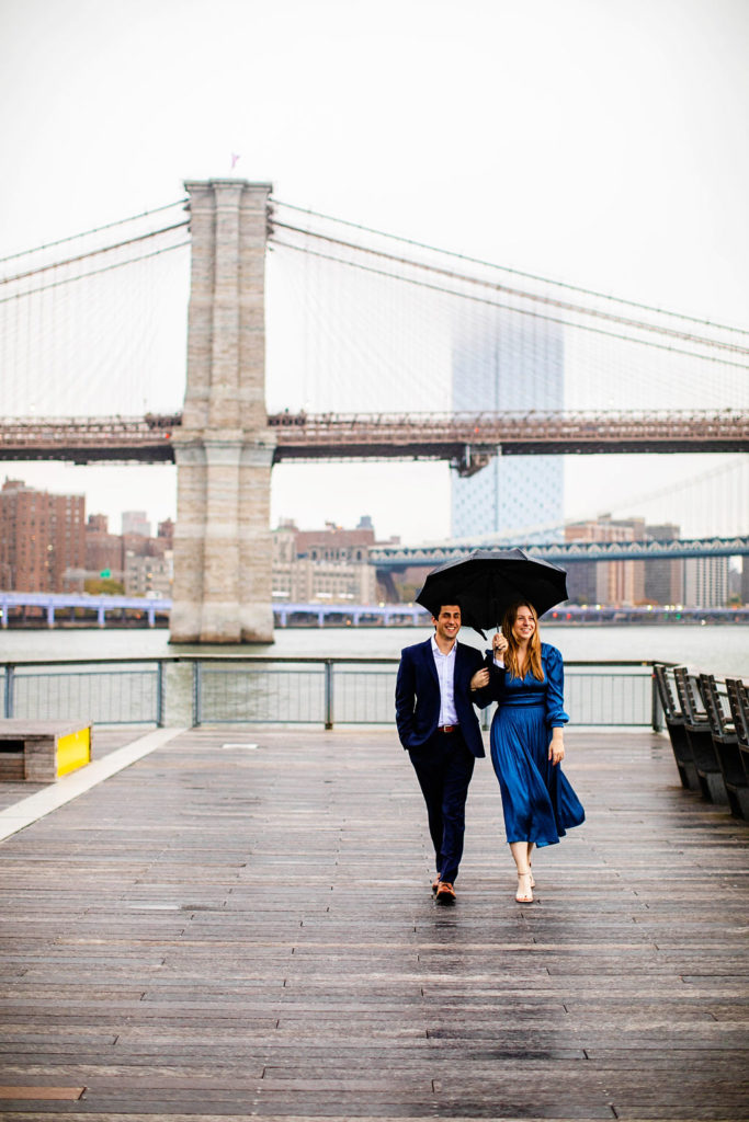 Couple walks umbrella with the Brooklyn Bridge in the background at their Manhattan engagement session at the South Street Seaport